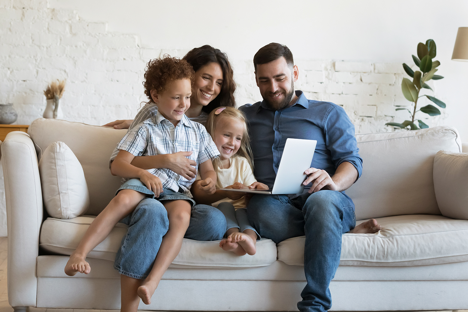 Happy caring parents holding little sibling kids in arms, resting on sofa, using laptop together, watching movie, interactive TV, talking on video call, chatting, laughing